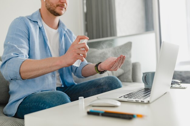 Feche as mãos do homem spray desinfetante anti-séptico no local de trabalho em casa trabalhando online no laptop