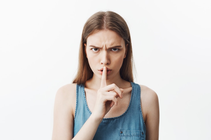 Foto grátis feche de bonita caucasiana jovem estudante com cabelos escuros na camisa azul, segurando a mão fromt dos lábios, fazendo silêncio gesto com expressão séria e insatisfeita. mulher tenta se acalmar l