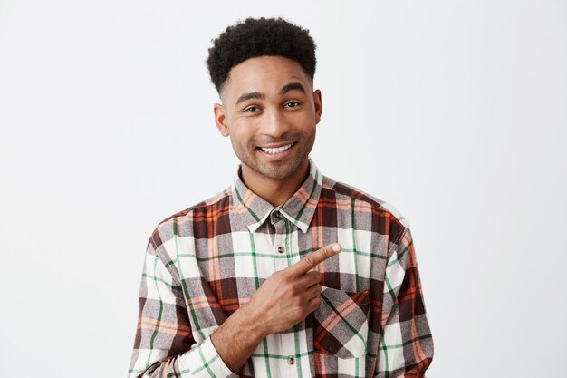 Feche de bonito alegre homem de pele bronzeada com penteado afro na camisa quadriculada casual, sorrindo brilhantemente, apontando de lado com o dedo indicador o parede branca. Copie o espaço