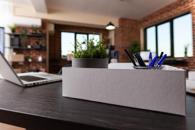 Feche de decorações de escritório em caixa e laptop na mesa. Planta de casa em vaso, suprimentos e instrumentos na mesa de madeira no espaço vazio de negócios. Vaso de flores e ornamentos modernos no trabalho