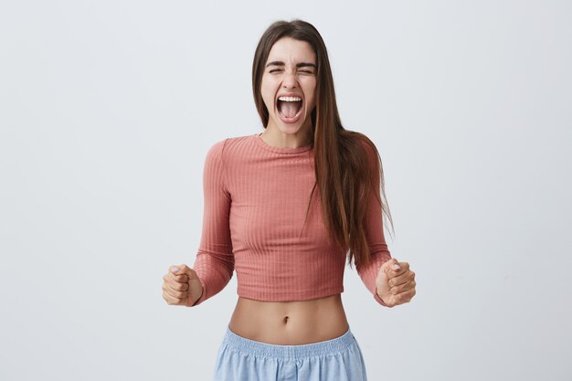 Feche de garota jovem estudante expressiva, com cabelos longos escuros na moda rosa top e shorts azuis gritando alto com os olhos fechados depois de ver aranha no banheiro.