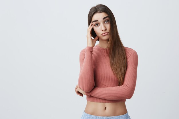 Feche de jovem atraente caucasiana infeliz com longos cabelos escuros na rosa elegante e esporte shorts segurando a cabeça com a mão com expressão cansada.