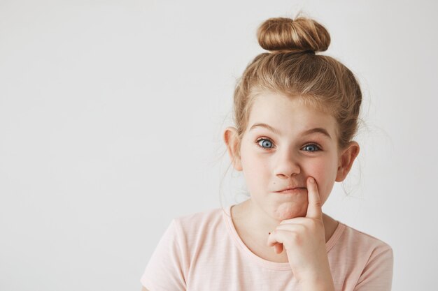 Feche de menina bonita com cabelo loiro no penteado de coque, segurando o dedo perto da boca, fazendo a expressão do rosto engraçado, se divertindo na sessão de fotos.