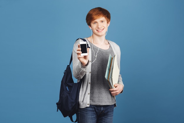 Feche de menino estudante ruivo lindo com mochila sorrindo, segurando muitos cadernos nas mãos, mostrando o telefone celular, ouvindo música favorita em fones de ouvido.