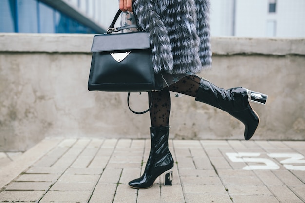 Feche detalhes de acessórios de uma mulher elegante andando pela cidade com um casaco de pele quente, inverno, clima frio, segurando uma bolsa de couro, pernas em botas, calçados, tendência da moda de rua