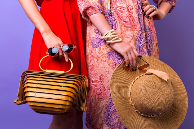 Feche detalhes de moda elegantes, duas mulheres com vestidos brilhantes posando em fundo roxo, segurando um chapéu de palha de óculos de sol e uma bolsa de madeira moderna super na moda.