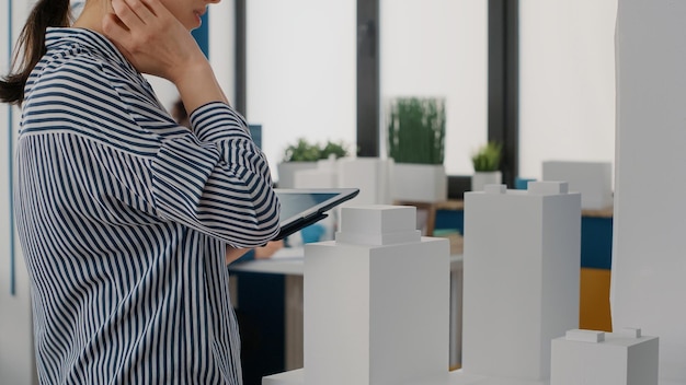 Foto grátis feche o arquiteto segurando o tablet e analisando o modelo de construção para planejar o layout. engenheiro de mulher usando dispositivo digital para projetar estrutura de construção e projeto de desenvolvimento.