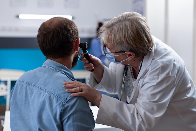 Feche o médico usando otoscópio para fazer consulta de ouvido com o paciente. Mulher otologista verificando infecção com instrumento de otorrinolaringologia na visita médica durante a pandemia de coronavírus.