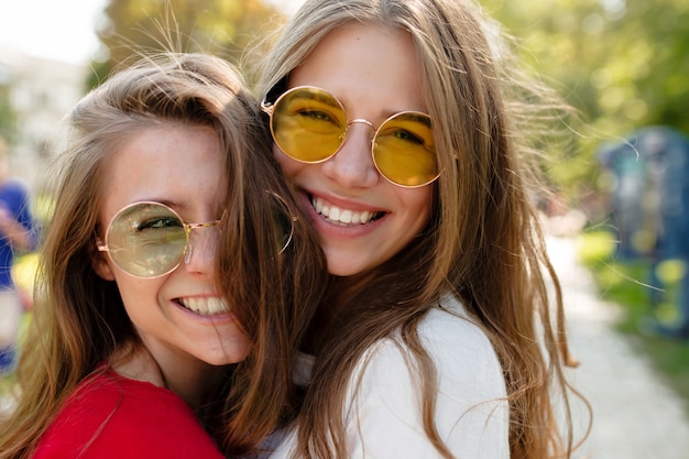 Feche o retrato ao ar livre de duas alegres melhores amigas de óculos brilhantes, sorrindo e posando no parque ensolarado. Duas amigas engraçadas passando um tempo livre comigo lá fora