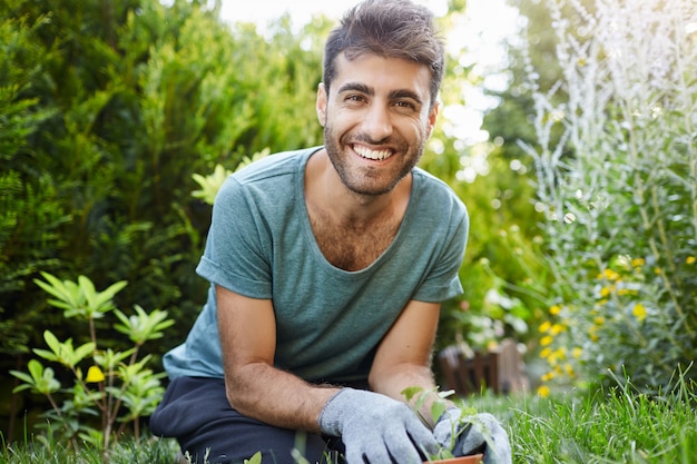 Foto grátis feche o retrato ao ar livre de uma jovem atraente barbudo caucasiano jardineiro masculino em t-shirt azul, sorrindo para a câmera, plantando sementes no jardim, regando as plantas.