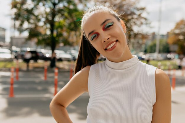 Feche o retrato ao ar livre de uma linda garota espetacular com maquiagem brilhante e cabelo coletado na camiseta está sorrindo sinceramente para a câmera no fundo da cidade