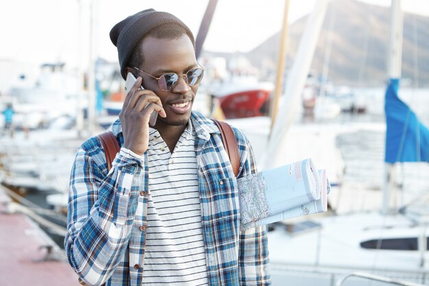 Feche o retrato ao ar livre do carismático jovem afro-americano em roupas elegantes, carregando o mapa de papel debaixo do braço, tendo uma boa conversa por telefone