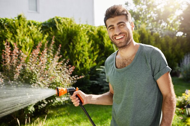 Feche o retrato ao ar livre do jovem bonito jardineiro masculino caucasiano sorrindo regando plantas, passando o verão em casa de campo.