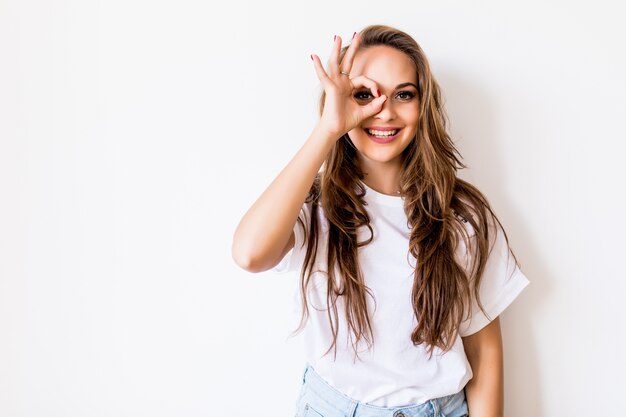 Feche o retrato da bela alegre mulher branca loira sorrindo, demonstrando os dentes brancos, olhando para a câmera por entre os dedos em um gesto bem. Expressões faciais, emoções e linguagem corporal