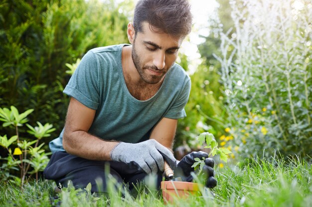 Feche o retrato da bela barbudo jardineiro masculino hispânico concentrado broto de plantio em vaso de flores com ferramentas de jardim, desfrutando de momentos de silêncio.