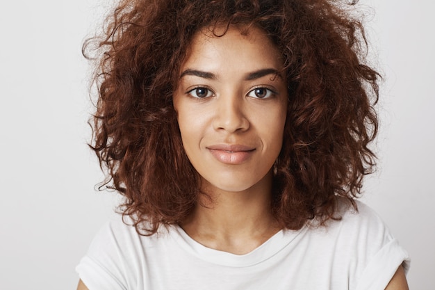 Feche o retrato da bela garota africana com olhos grandes, sorrindo com um sorriso, sentindo-se confiante e calmo, sendo atraente e atraente.