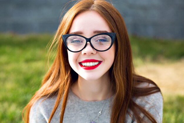 Feche o retrato da deslumbrante linda menina adolescente ruiva, sorriso incrível, lábios vermelhos, cabelos longos, óculos de sol vintage claros, humor positivo.