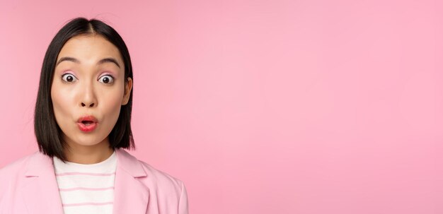 Feche o retrato da empresária asiática corporativa olhando surpresa e espantada com a câmera em pé de terno contra o fundo rosa