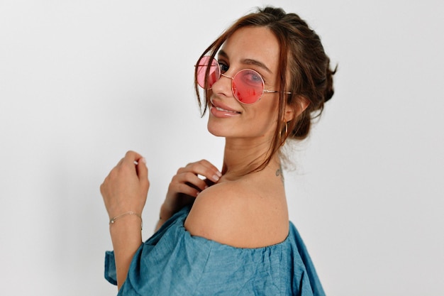 Feche o retrato da encantadora garota bronzeada europeia em óculos rosa redondos posando para a câmera de volta com um lindo sorriso sobre fundo isolado