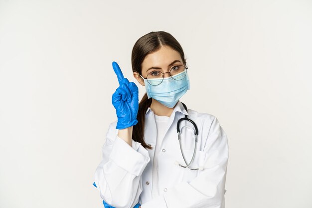 Feche o retrato da médica em óculos, máscara facial médica e luvas, apontando o dedo para cima...