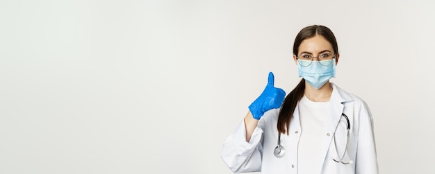 Feche o retrato da médica médica com máscara facial de coronavírus mostrando os polegares para cima e sorrindo