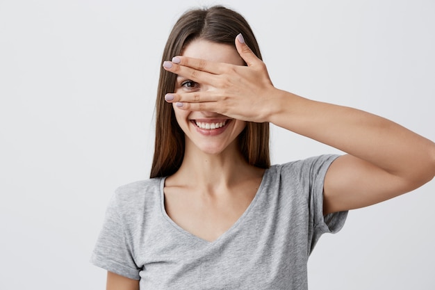 Feche o retrato da menina alegre bonita jovem estudante caucasiano com cabelos longos escuros na camiseta cinza na moda sorrindo com dentes, olhos de roupas com a mão, olhando pelo dedo com um olho.