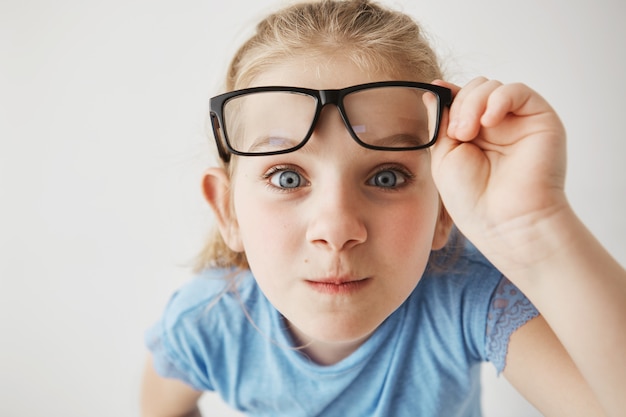 Foto grátis feche o retrato da menina curiosa com grandes olhos azuis em pé perto e, segurando os óculos com a mão.