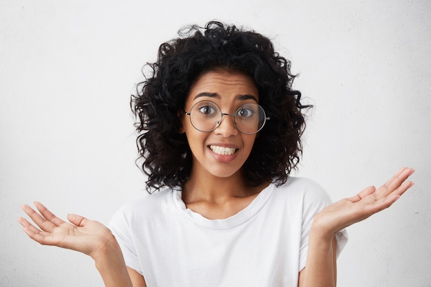 Foto grátis feche o retrato da mulher afro-americana vestindo camiseta branca, sentindo-se culpado, confuso, fazendo um gesto impotente com as mãos, tendo opa expressão no rosto. emoções e sentimentos humanos