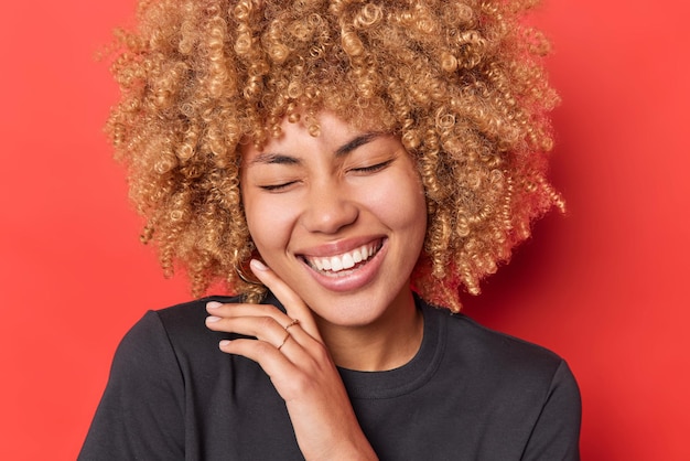 Feche o retrato da mulher de cabelos cacheados toca o rosto suavemente fecha os olhos do prazer goza de suavidade de sorrisos de pele mostra amplamente os dentes brancos isolados sobre fundo vermelho vívido. emoções sinceras