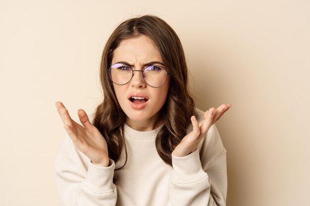 Feche o retrato da mulher de óculos parecendo confuso e frustrado, não consigo entender algo estranho, de pé sobre fundo bege.