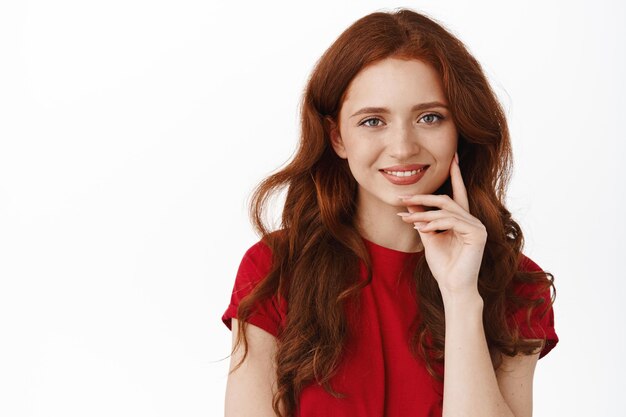 Feche o retrato da mulher ruiva autêntica, sorrindo e tocando o rosto natural sem maquiagem, olhando feliz e fofo para a câmera, de pé na camiseta vermelha contra fundo branco.