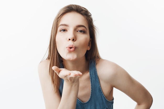 Feche o retrato de alegre jovem bela mulher caucasiana, com cabelos escuros na camisa azul, soprando ar beijo