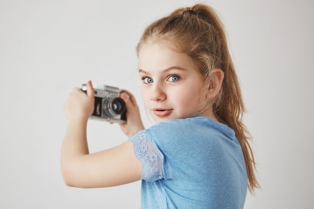 Feche o retrato de alegre linda garota com cabelos loiros e olhos azuis, com expressão interessada, vai tirar uma selfie.