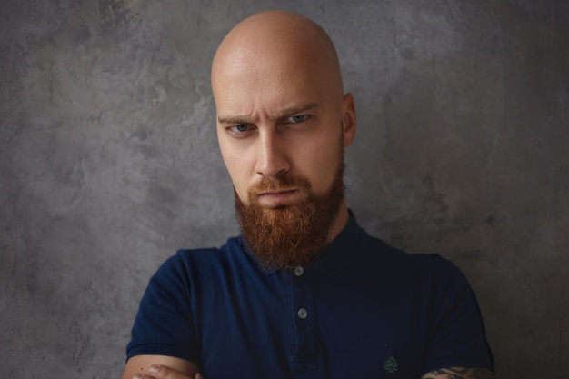 Feche o retrato de bonito carrancudo jovem careca com barba espessa, tendo olhar carrancudo e carrancudo com os olhos cheios de raiva e raiva. Expressões faciais e emoções humanas negativas