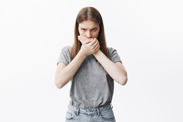 Feche o retrato de infeliz erótica menina de cabelos escuros com cabelos longos em roupas da moda fechando a boca com as duas mãos, com expressão de culpa depois de dizer algo ofensivo