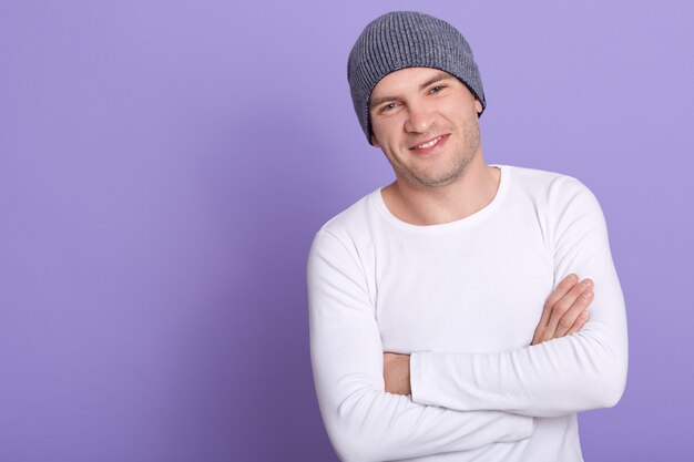 Feche o retrato de jovem atraente vestindo camisa branca casual manga longa e boné cinza, posando isolado no lilás, em pé com os braços cruzados. Copie o espaço.