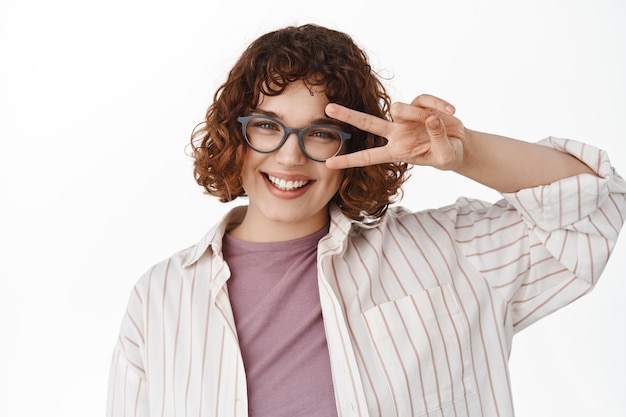 Feche o retrato de sorridente jovem natural com cabelos cacheados, usando óculos, mostrando paz kawaii v-sign perto do olho, parecendo feliz e positivo para a câmera, fundo branco.