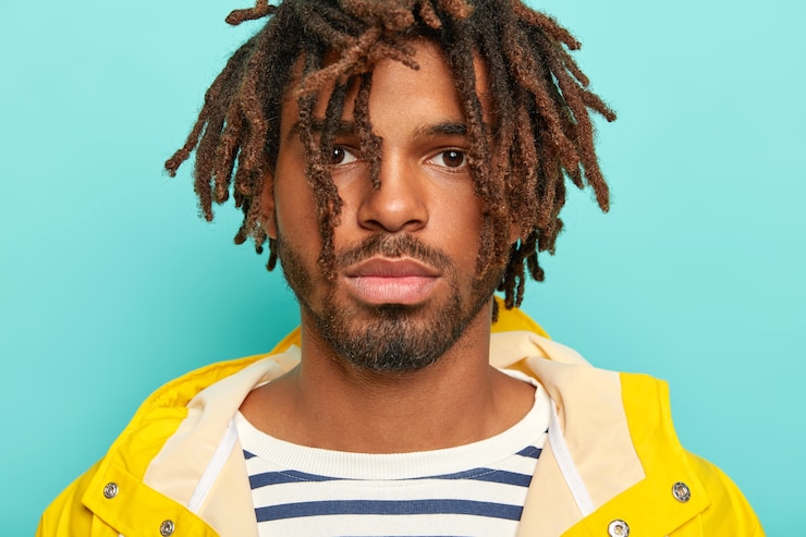 Foto grátis feche o retrato de um hipster sério de pele escura com drealocks, tem lábios carnudos, barba e bigode, usa um macacão listrado e capa de chuva amarela, escuta com atenção interlocutor, isolado na parede azul