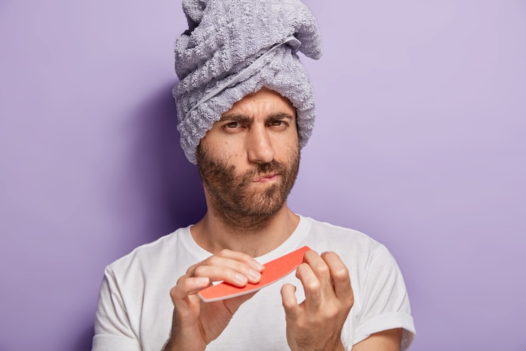 Foto grátis feche o retrato de um homem sério com a barba por fazer com uma toalha macia na cabeça, faz as unhas depois de tomar banho, usa lixa de unha, se preocupa com sua beleza, usa camiseta branca, isolado na parede roxa