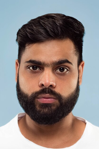 Feche o retrato de um jovem hindu com barba na camisa branca, isolada na parede azul. Emoções humanas, expressão facial, conceito de anúncio. Espaço negativo. De pé e parecendo calmo.