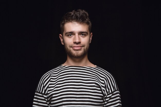 Feche o retrato de um jovem isolado no fundo preto do estúdio. Photoshot de emoções reais do modelo masculino. Sorrindo, me sentindo feliz. Expressão facial, conceito de emoções humanas puras e claras.