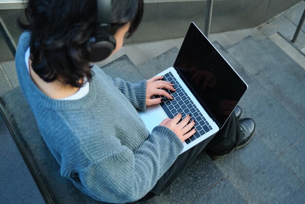 Feche o retrato de uma aluna que trabalha no laptop e ouve música em fones de ouvido, tela de computador em branco