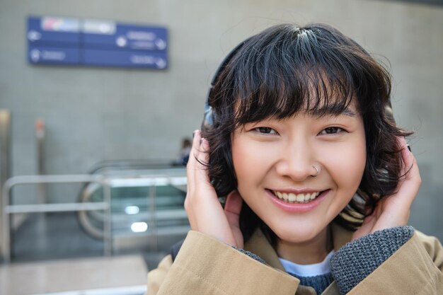 Feche o retrato de uma elegante garota coreana parada na rua ouvindo música em fones de ouvido e sorrindo