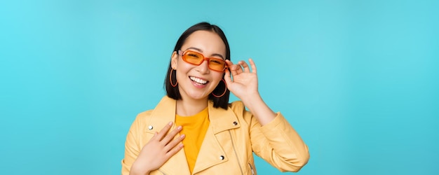 Feche o retrato de uma elegante mulher asiática em óculos de sol rindo e sorrindo parecendo feliz posando em roupas da moda sobre fundo azul