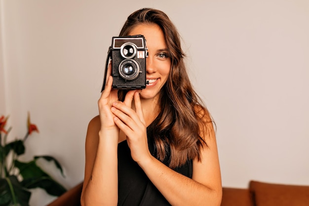 Feche o retrato de uma encantadora garota elegante com longos cabelos escuros está segurando uma câmera retrô e sorrindo sobre fundo bege