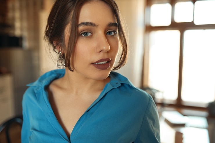 Foto grátis feche o retrato de uma garota atraente glamourosa com olhos azuis e cabelo recolhido posando dentro de casa, contra o fundo da janela, usando batom marrom e camisa azul. conceito de pessoas e estilo de vida