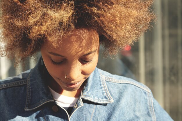 Feche o retrato de uma garota morena hippie com corte de cabelo afro e piercing no nariz, vestida com uma jaqueta jeans da moda, posando ao ar livre com os olhos fechados e um sorriso fofo, aproveitando o bom tempo durante uma caminhada