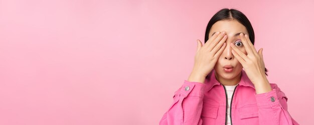 Feche o retrato de uma jovem asiática que parece surpresa expressa surpresa e maravilha espiando por entre os dedos em pé sobre fundo rosa