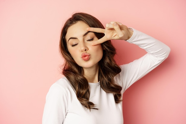 Feche o retrato de uma jovem coquete sorrindo, mostrando paz, gesto de v-sign e posando feliz, em pé contra um fundo rosa