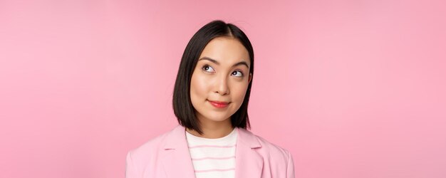 Feche o retrato de uma jovem empresária asiática pensando sorrindo pensativa e olhando para o canto superior esquerdo sobre fundo rosa